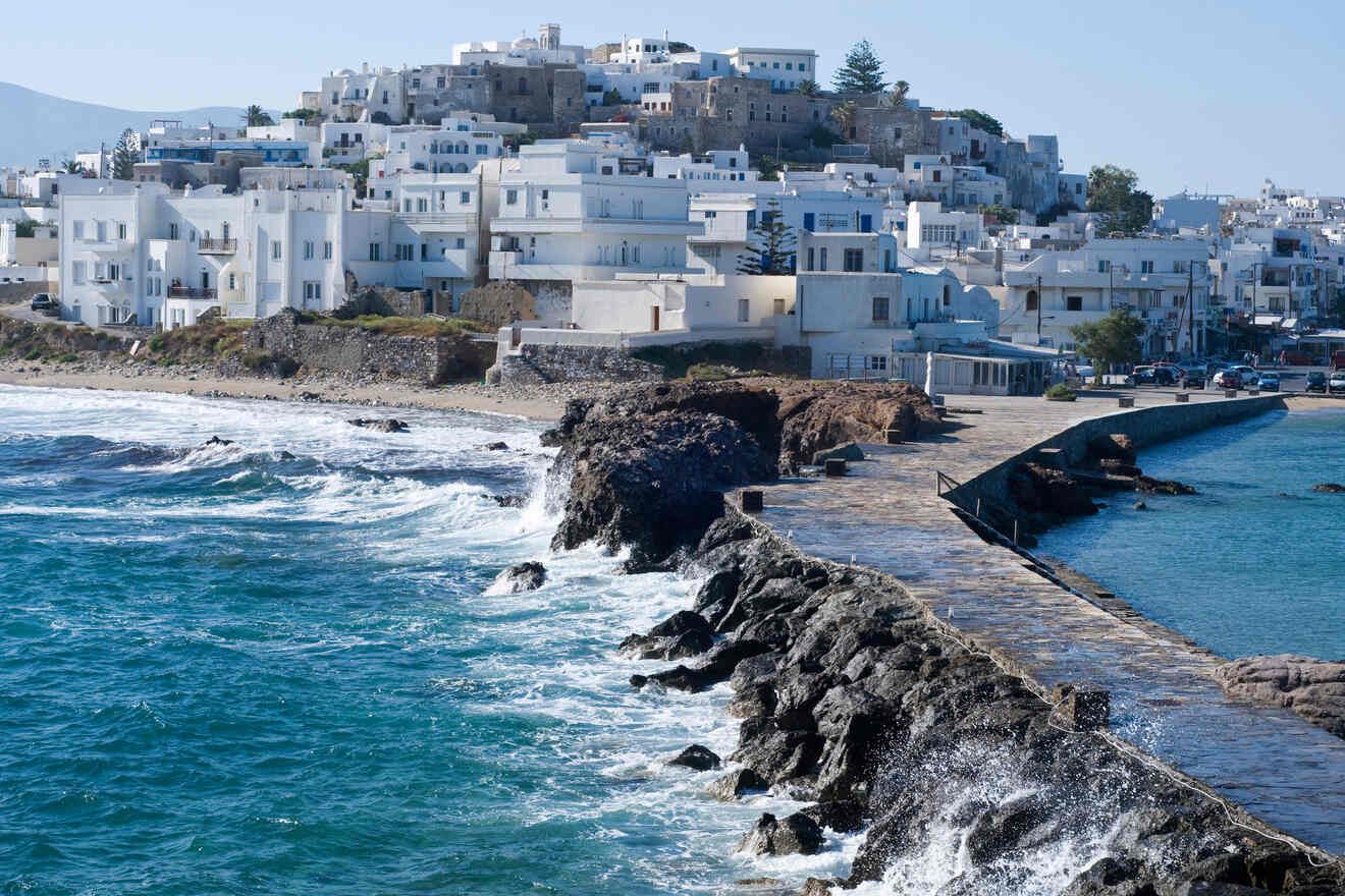 Coastal view of the whitewashed town of Naxos near the rocky shore with waves breaking, under a clear blue sky.
